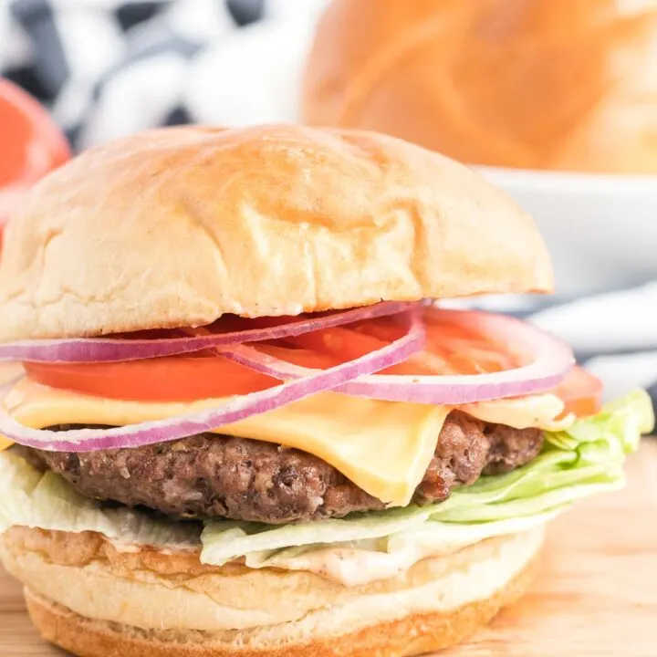 Hamburger on a wooden board topped with sauce, lettuce, cheese, tomato and red onions.