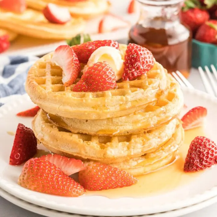 Stack of four cooked waffles with butter, strawberries and maple syrup.