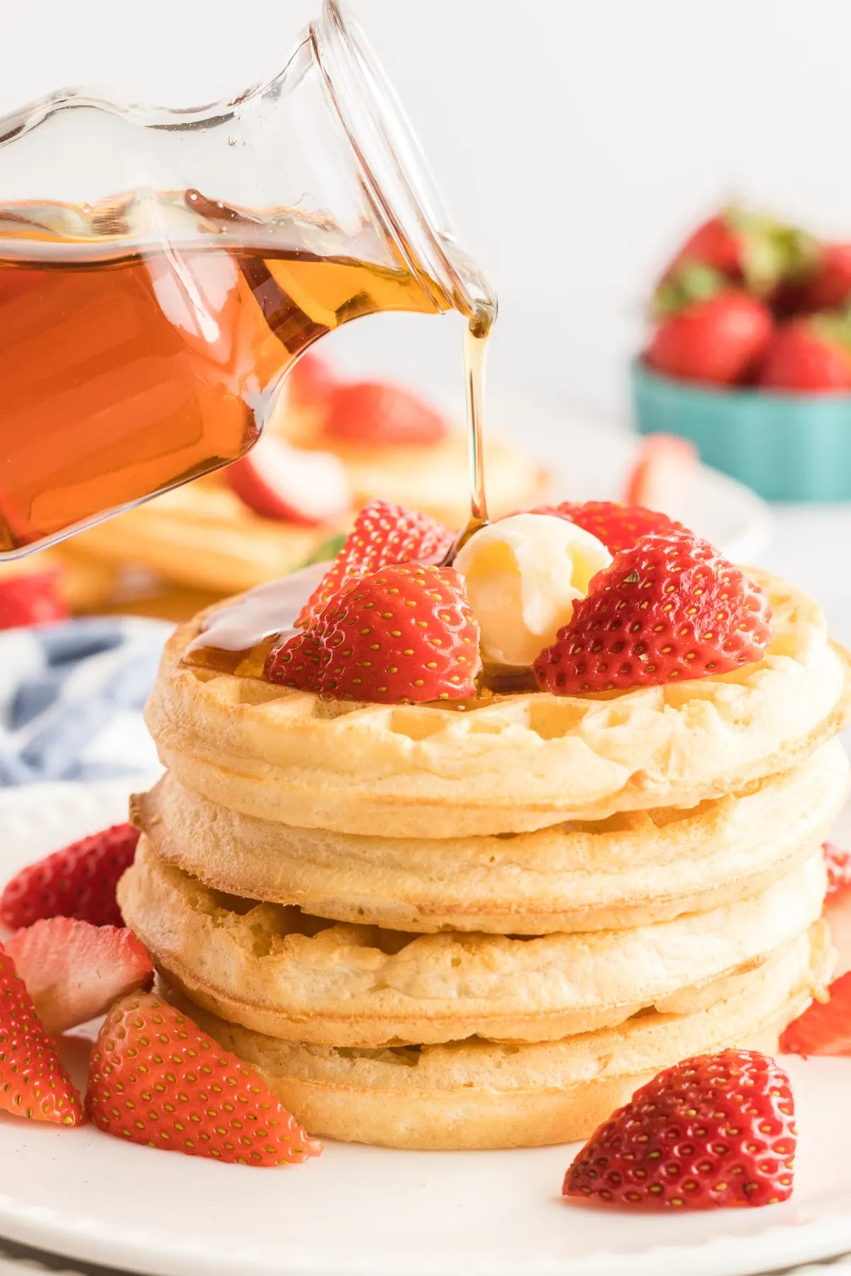 Four waffles in a stack with strawberries a jar of maple syrup being drizzled on top. 