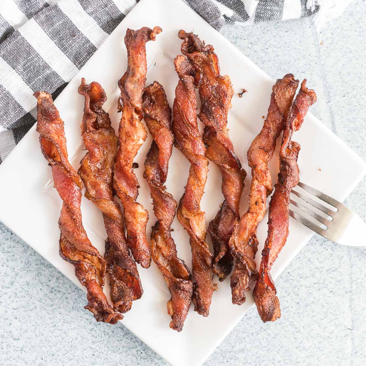 Cooked twisted bacon on a square white plate with a fork.