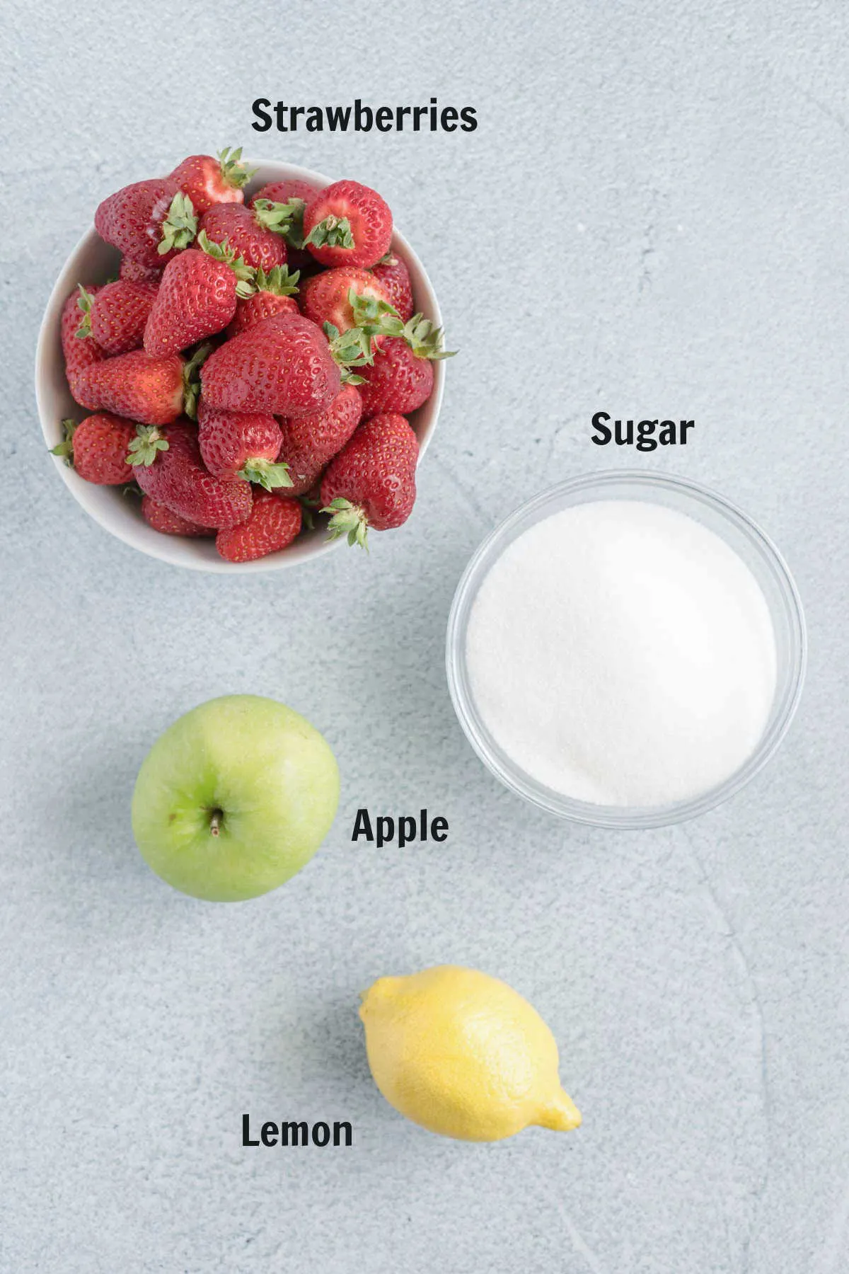 A bowl of fresh strawberries, a bowl of granulated sugar, a Granny Smith apple and a fresh lemon.