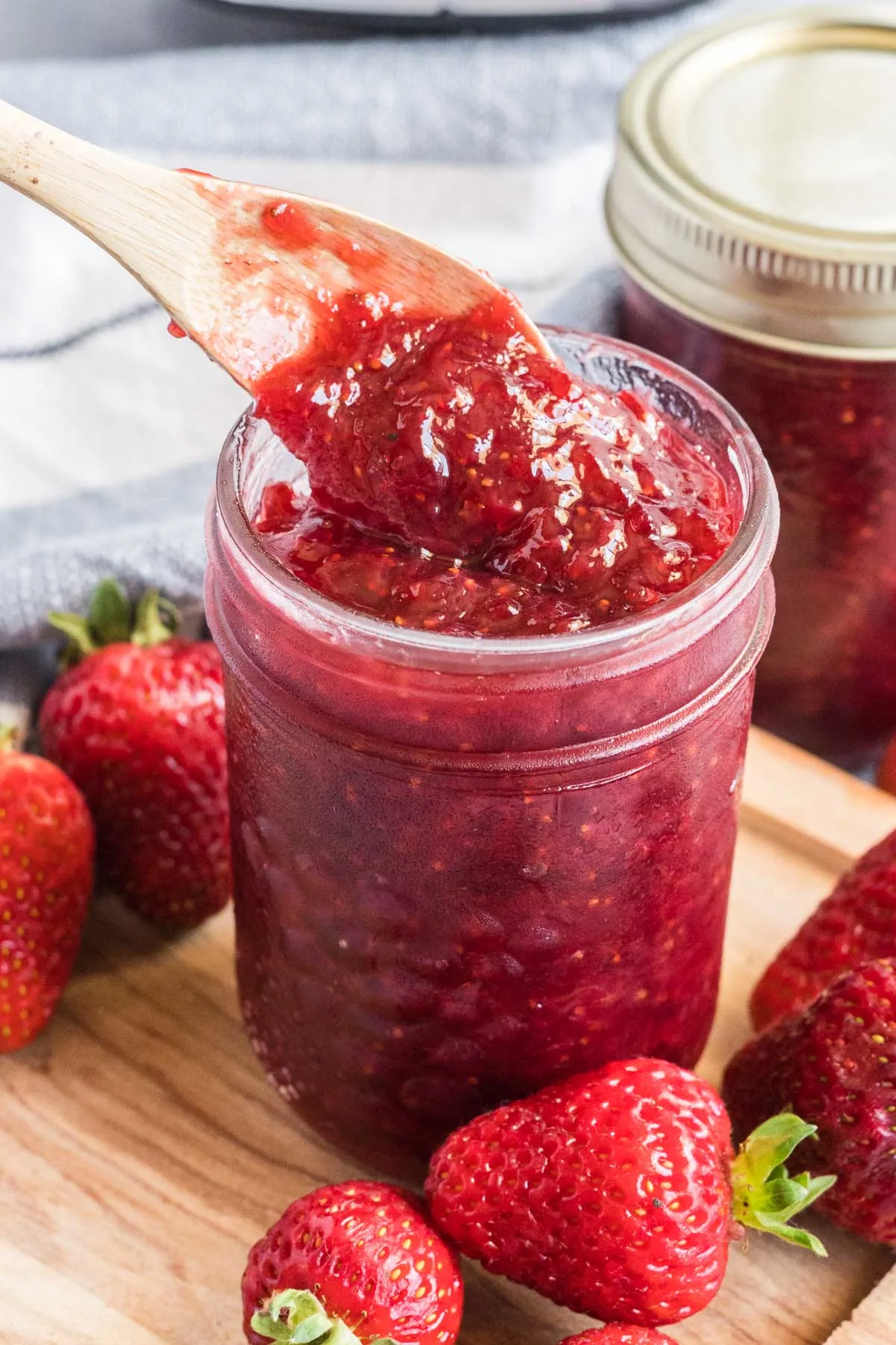 Strawberry jam being spooned out of a small mason jar. Fresh strawberries scattered around the jar.