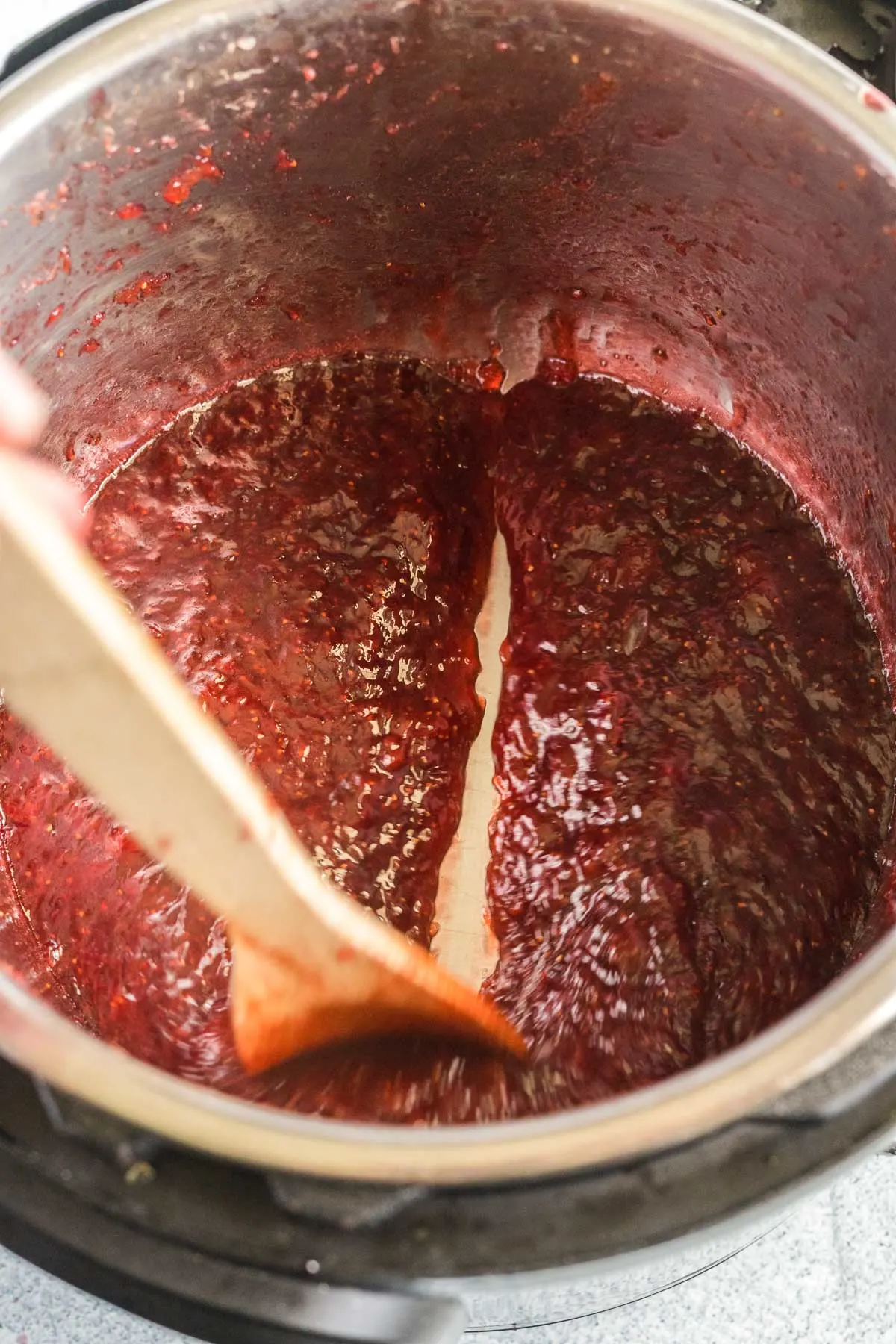 A wooden spoon being pulled through cooked jam in an instant pot so you can see the bottom of the pot.