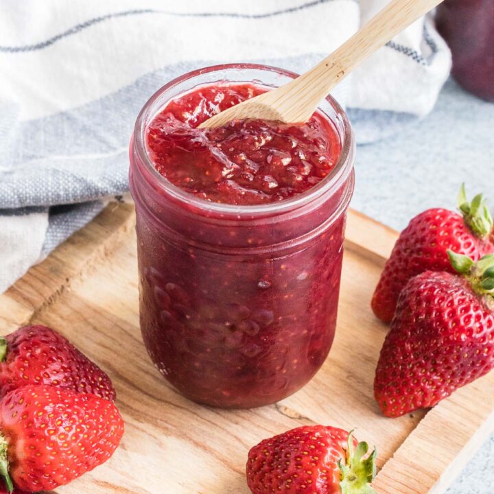 Strawberry jam in a small mason jar with a spoon. Fresh strawberries scattered around the jar.