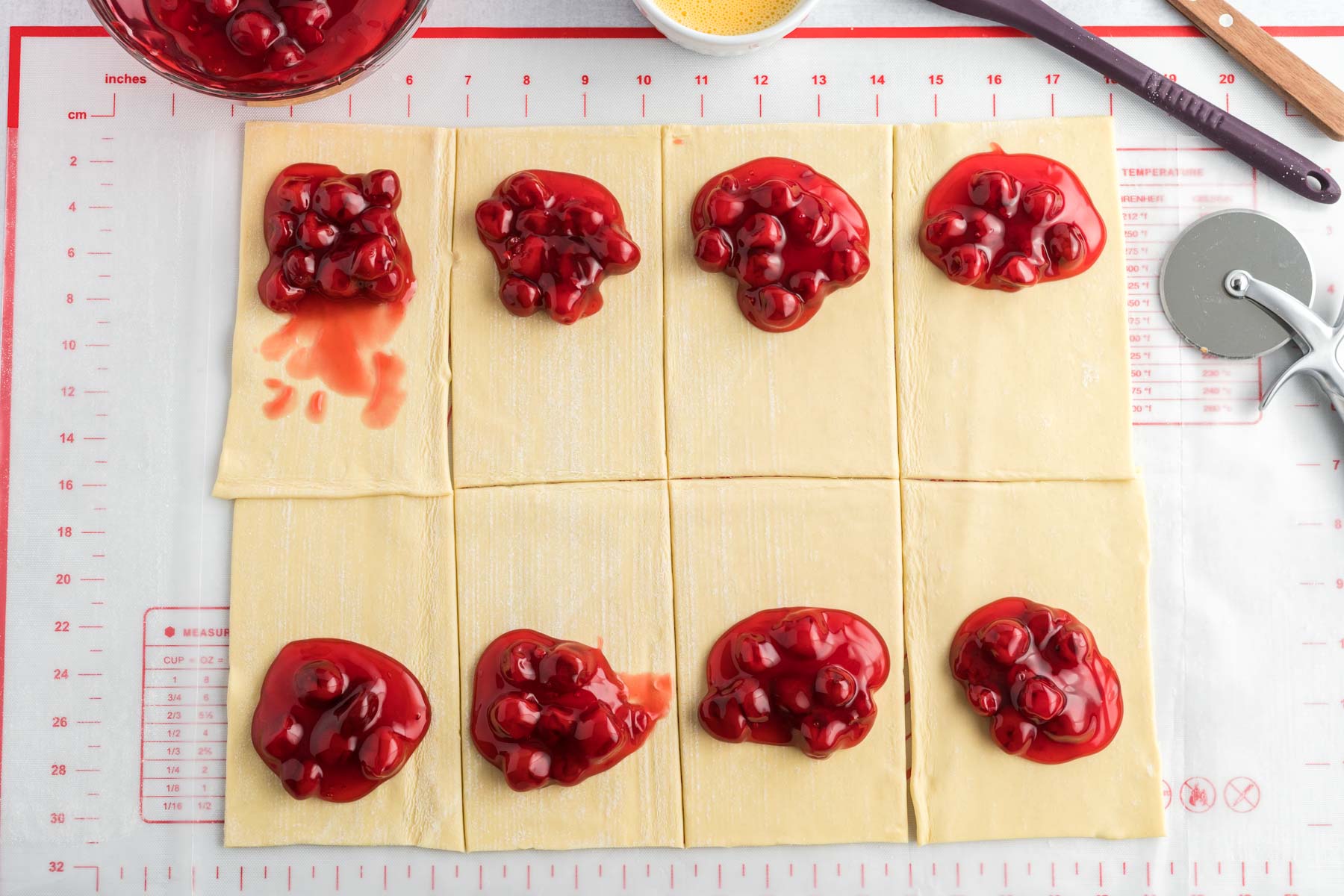 A sheet of puff pastry cut into 8 squares with a dollop of cherry pie filling on each square.