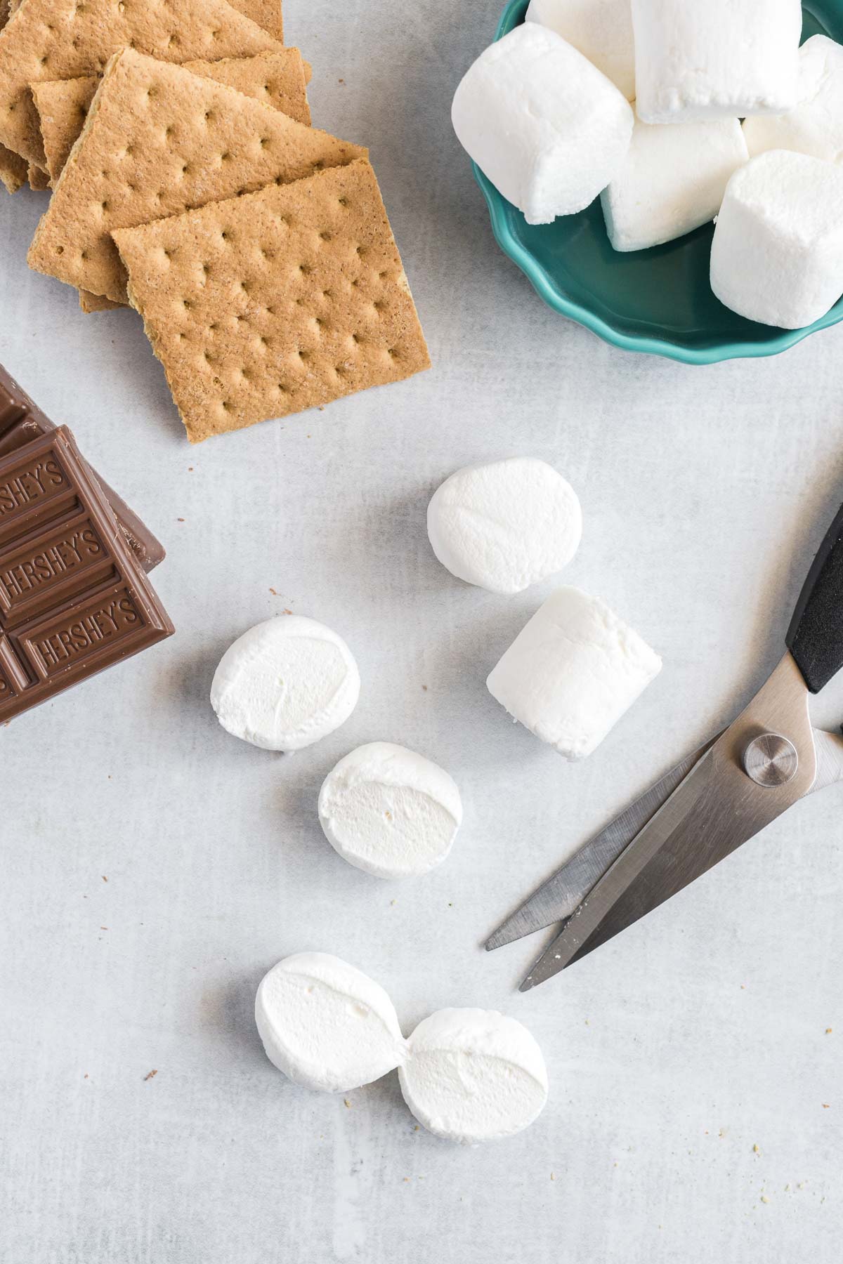 A stack of graham crackers and a bowl of marshmallows. Two marshmallows are cut in half and kitchen scissors are beside them.