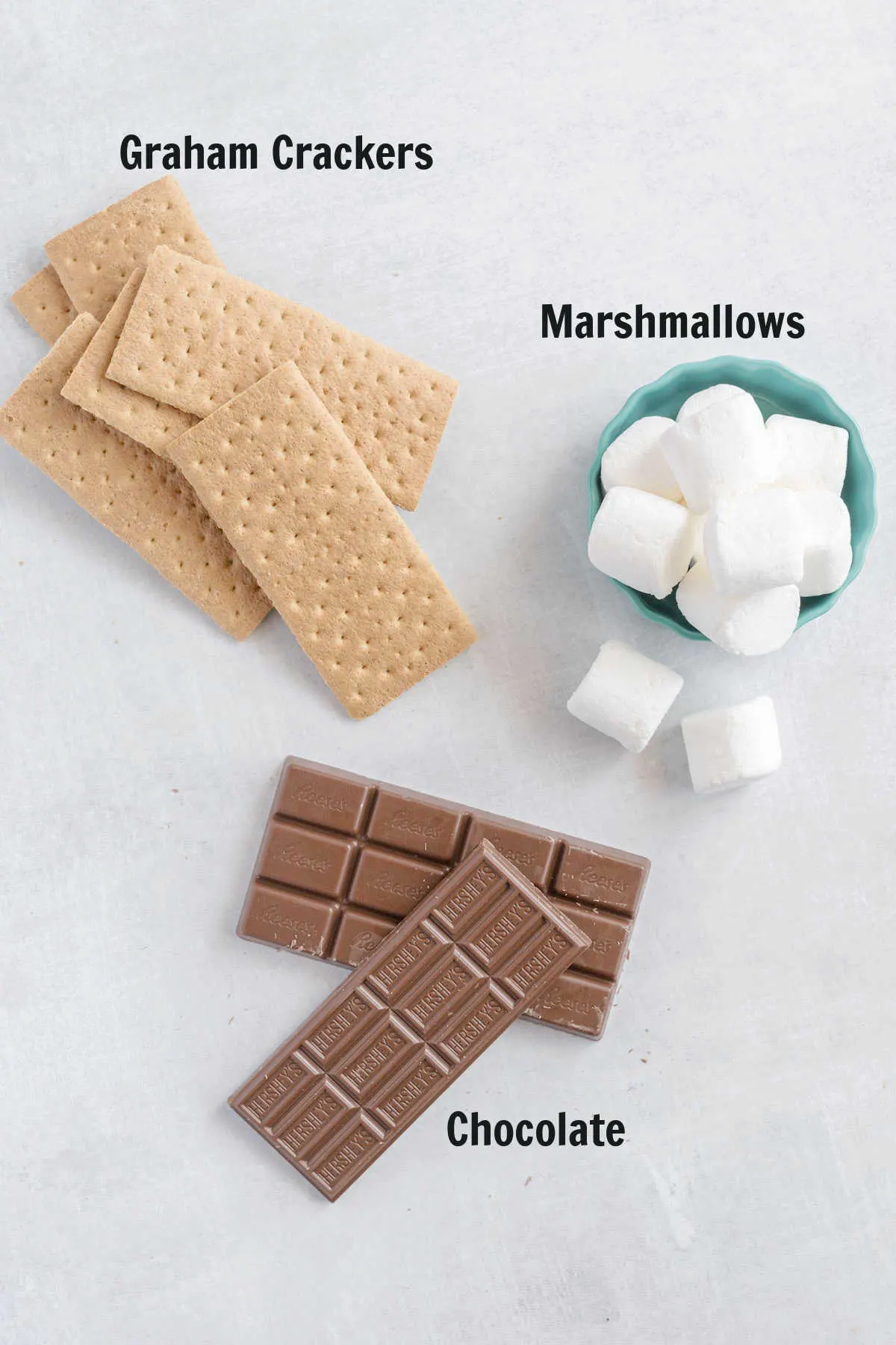 A stack of graham crackers, two chocolate bars and a bowl of large marshmallows.