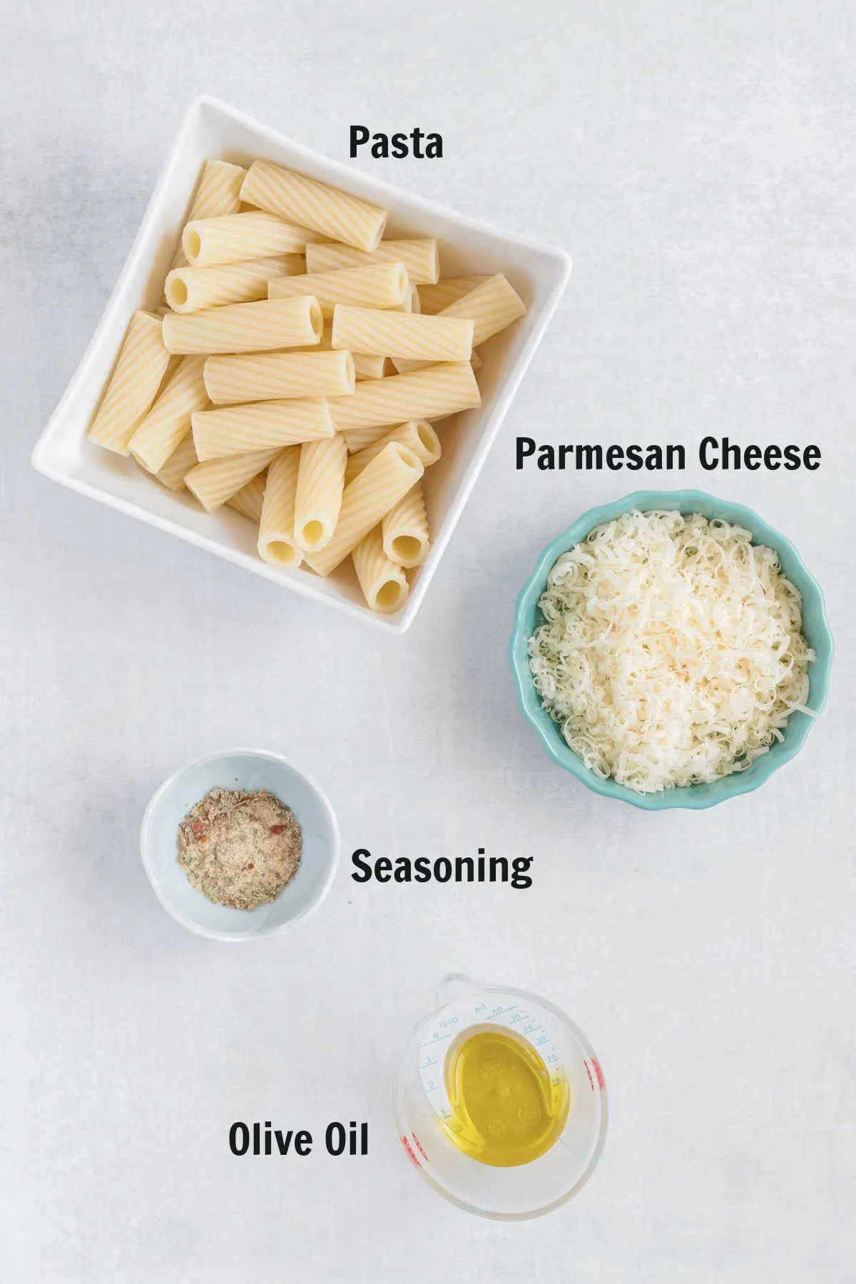 A bowl of cooked rigatoni, a dish of shredded parmesan, all purpose seasoning, and olive oil. 