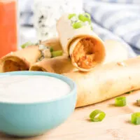 Chicken taquitos topped with crumbled blue cheese and green onion with a dish of blue cheese dip.