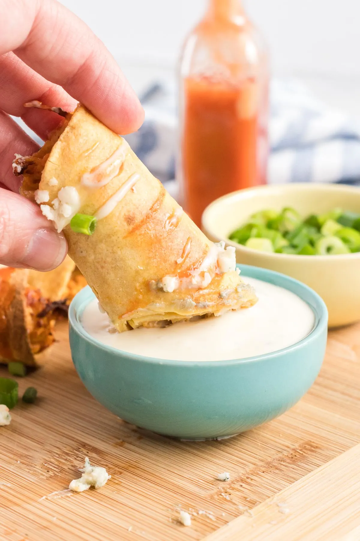 Taquito being dipped in blue cheese and a bottle of hot sauce in the background.