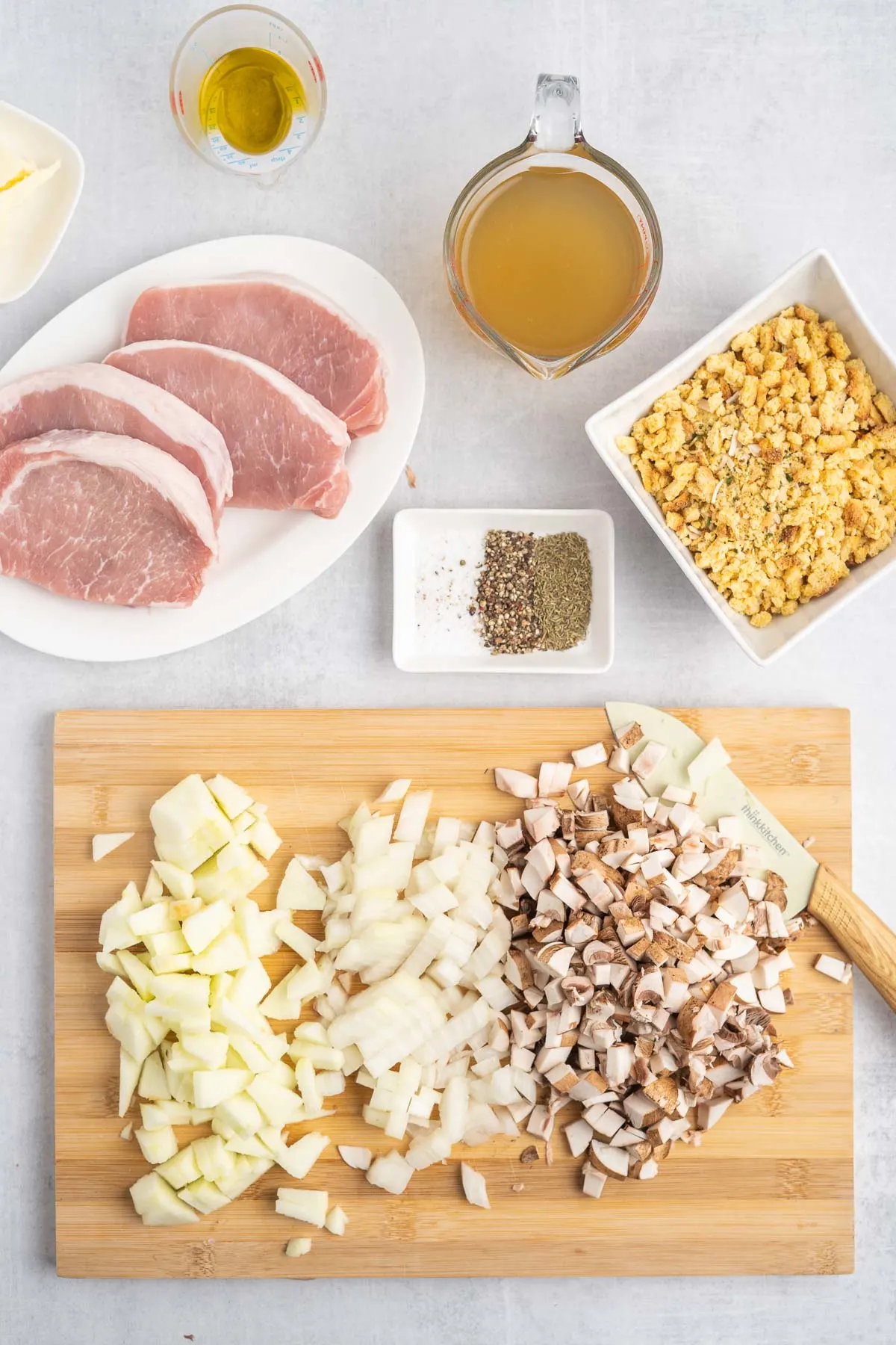 Chopped ingredients on a cutting board with a plate of boneless pork chops and a bowl of stuffing mix. 