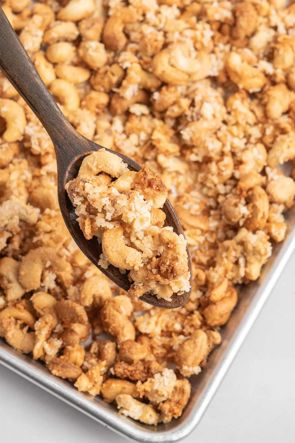 Coconut coated cashews on a baking sheet with a spoonful on a wooden spoon. 