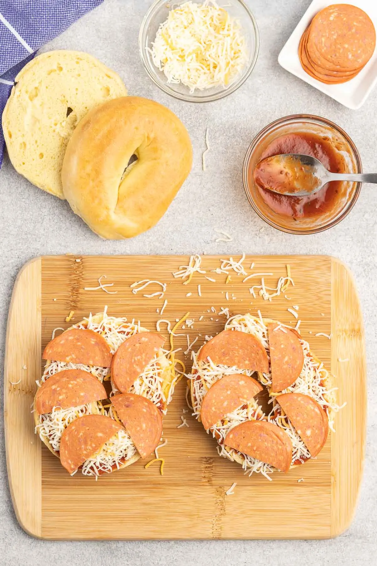 Two bagel halves on a cutting board topped with pizza sauce, cheese and pepperoni slices.
