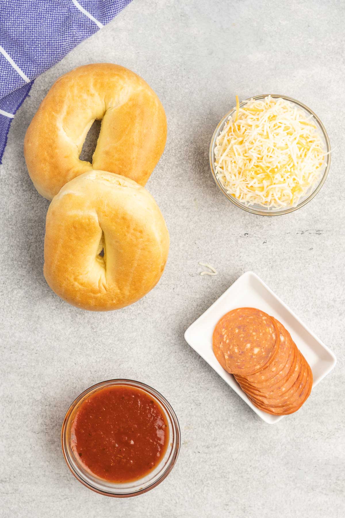 Two bagels and a dishes of pizza sauce, shredded cheese and pepperoni slices. 