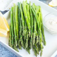 Roasted asparagus on a platter with garlic dip and lemon wedges.