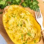 Half of a cooked spaghetti squash on a cutting board with the flesh shredded, garnished with fresh parsley.