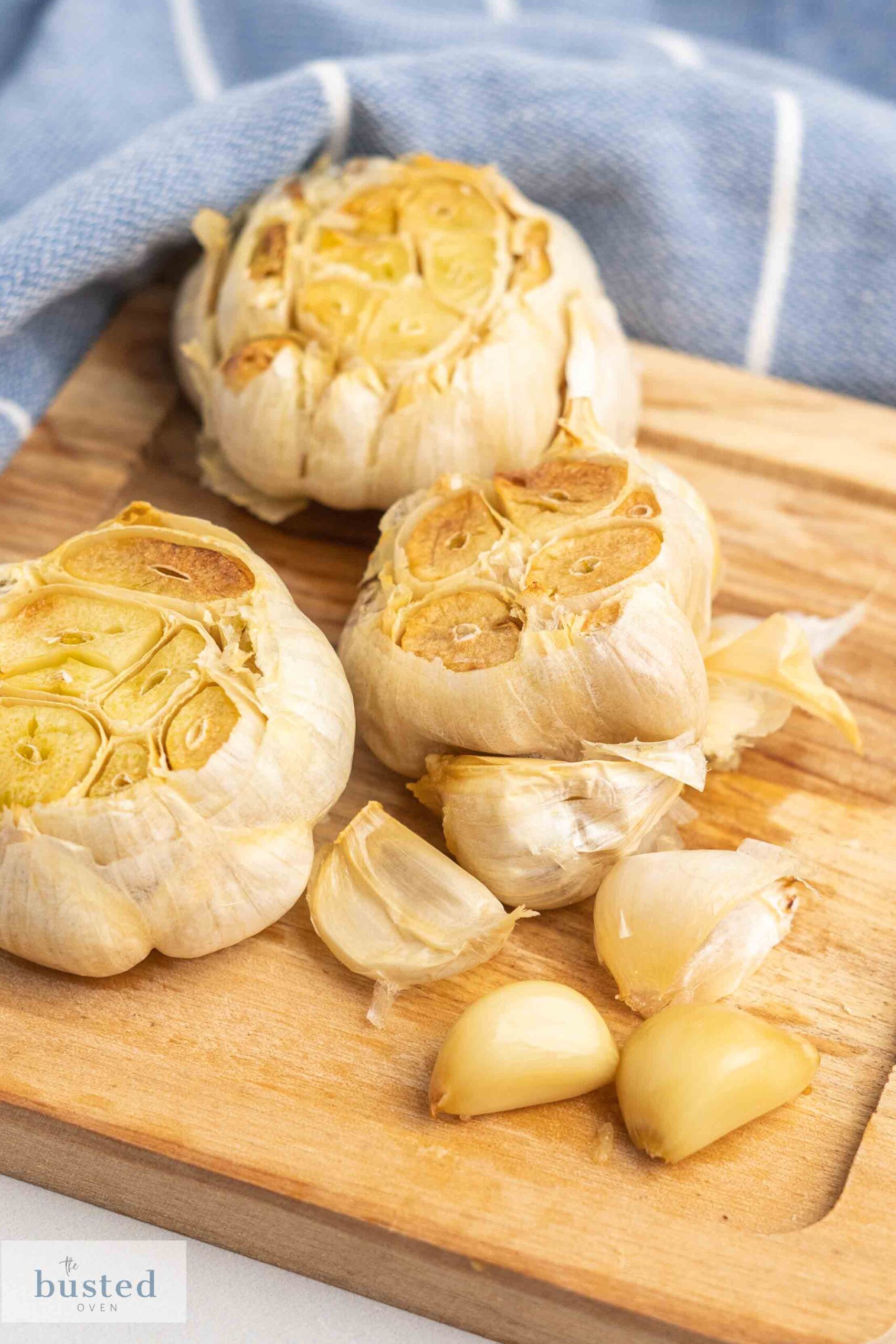 Three roasted garlic bulbs on a wooden plate and a few peeled garlic cloves. 