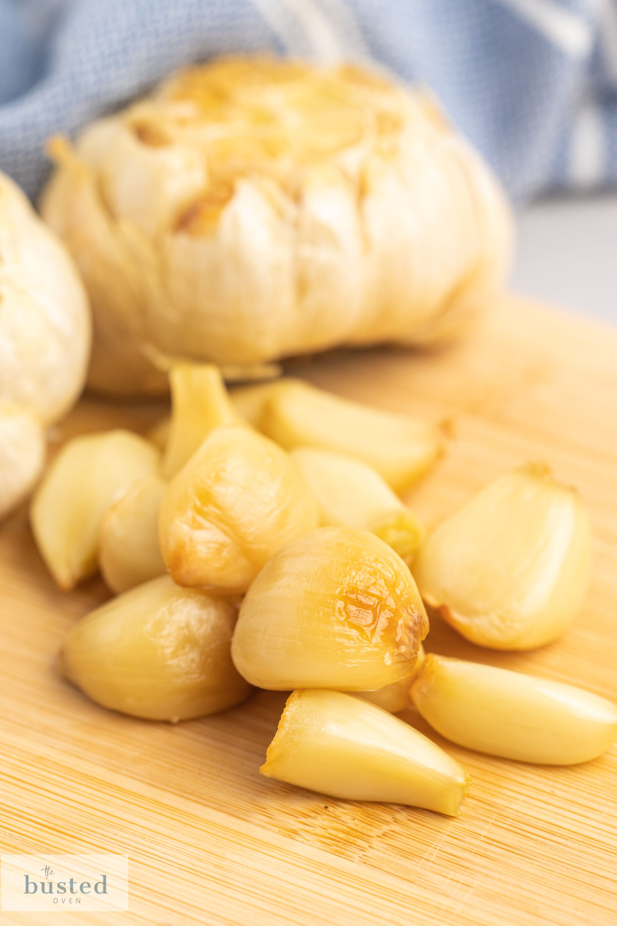 Peeled roasted garlic cloves on a wooden board and garlic bulbs in the background. 