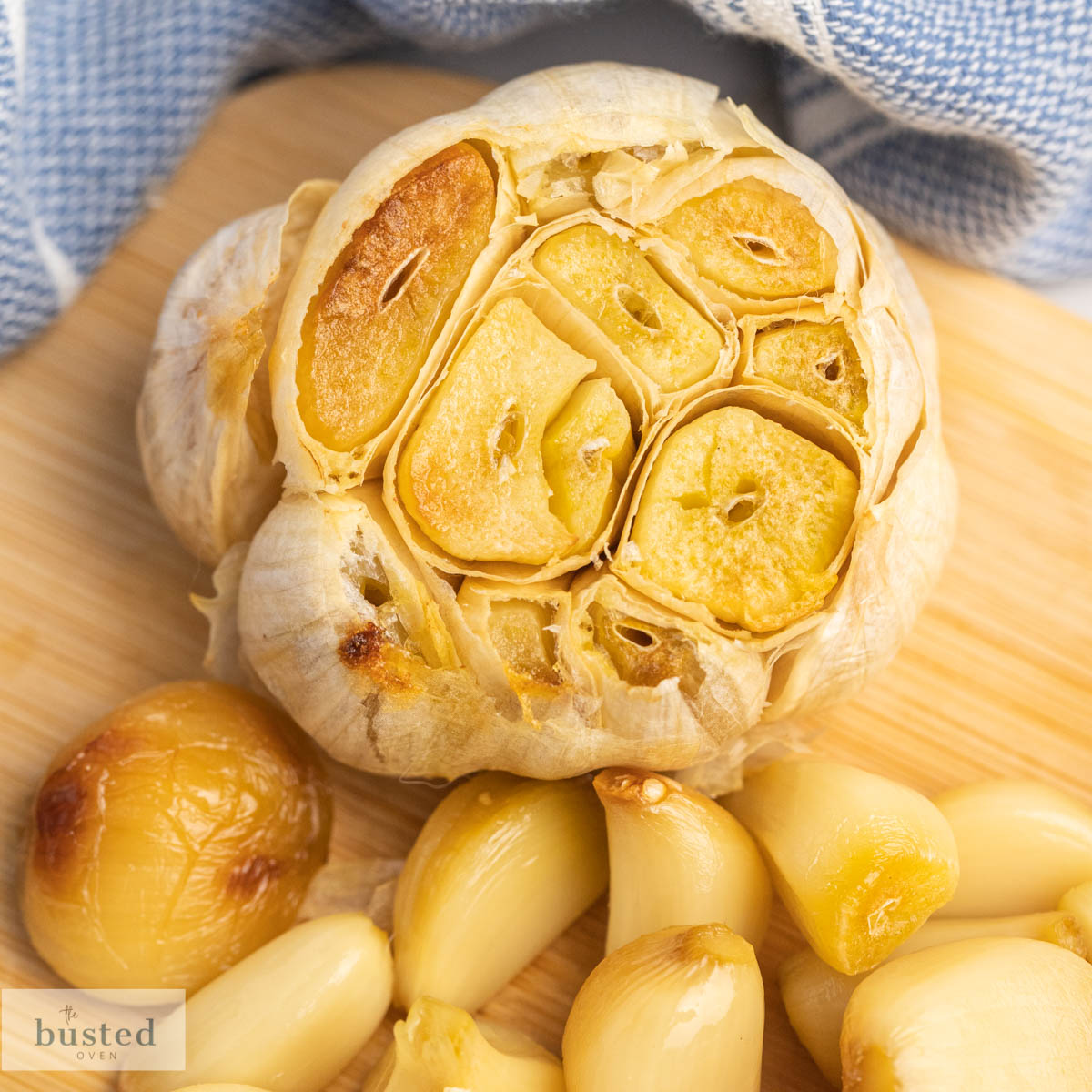 A bulb of roasted garlic on a small wooden cutting board and a pile of peeled roasted garlic cloves. 