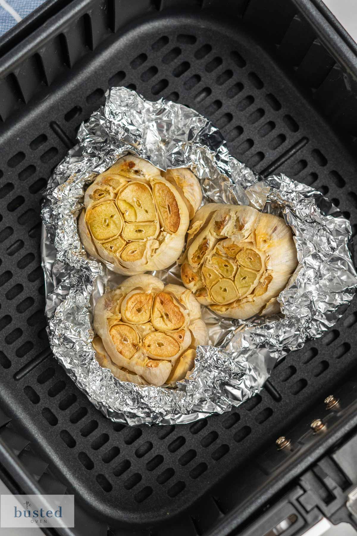 Three garlic bulbs with top cut off wrapped in tinfoil in an air fryer basket. 