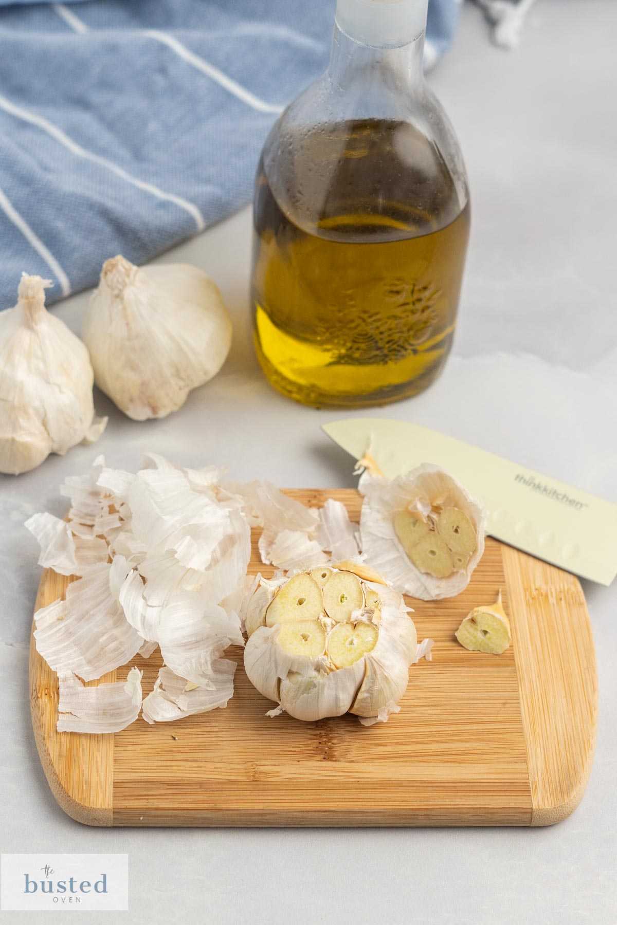 Garlic bulb on a cutting board with the top cut off and a bottle of olive oil beside it. 