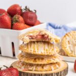 Four strawberry hand pies stacked on a small wooden plate with a basket of strawberries in the background.