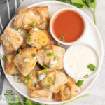 A plate of crispy wontons garnished with crumbled blue cheese and sliced green onion along side dishes of hot sauce and blue cheese dip.